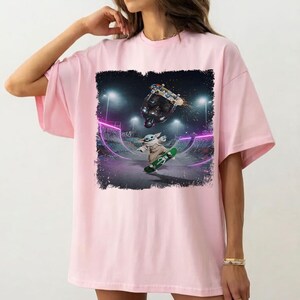 K&ouml;nnte beinhalten: Hellrosa T-Shirt mit einem Aufdruck eines Charakters, der in einem Skatepark skateboardet. Der Aufdruck zeigt ein gr&uuml;nes Skateboard, eine Figur und einen schwarzen Skateboarder. Der Hintergrund zeigt einen Skatepark mit neonvioletten Lichtern.