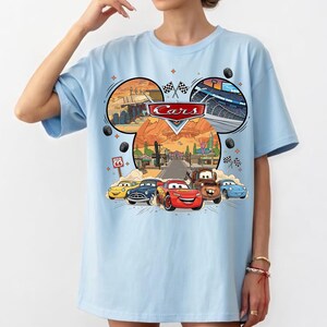 May include: Light blue t-shirt featuring a colorful graphic of the Disney Pixar Cars movie. The design includes the Cars logo, race tracks, and images of the characters Lightning McQueen, Mater, and others. The shirt has short sleeves.