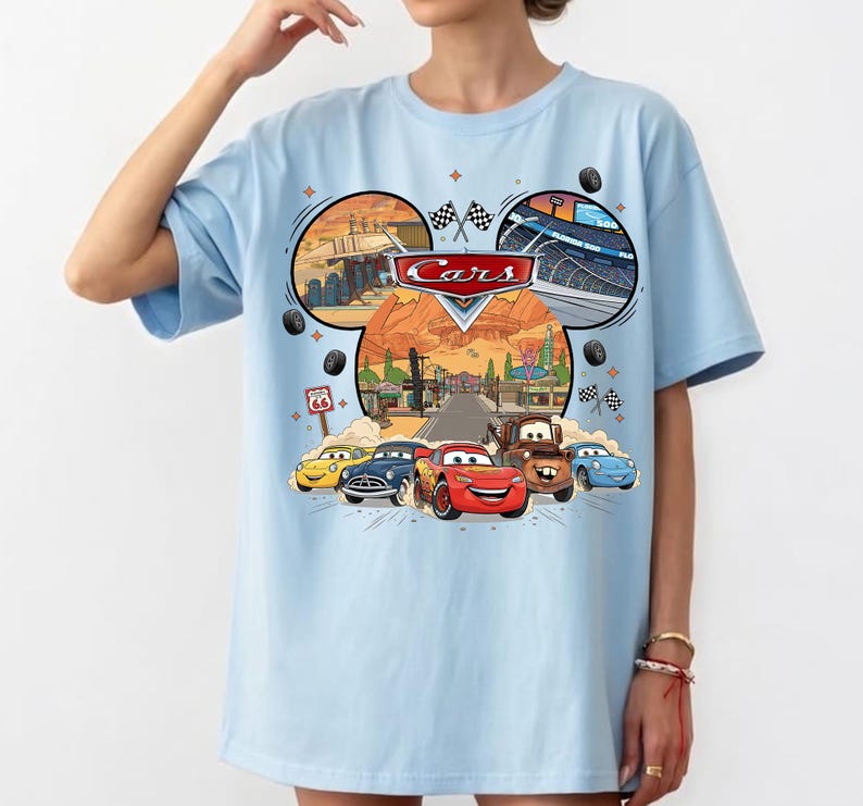 May include: Light blue t-shirt featuring a colorful graphic of the Disney Pixar Cars movie. The design includes the Cars logo, race tracks, and images of the characters Lightning McQueen, Mater, and others. The shirt has short sleeves.