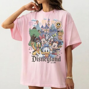 Op de afbeelding: Een lichtroze t-shirt met een kleurrijke grafische print van Disney-figuren waaronder Mickey Mouse, Minnie Mouse, Donald Duck, Goofy en Pluto voor een kasteel, met de tekst Disneyland Est. 1955 onder de figuren gedrukt.