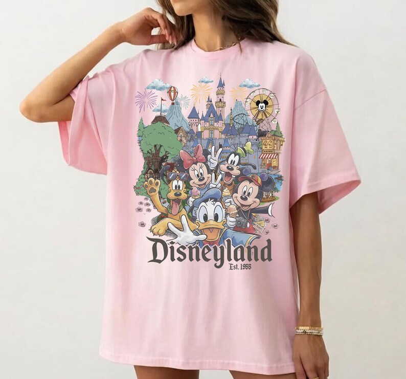 Op de afbeelding: Een lichtroze t-shirt met een kleurrijke grafische print van Disney-figuren waaronder Mickey Mouse, Minnie Mouse, Donald Duck, Goofy en Pluto voor een kasteel, met de tekst Disneyland Est. 1955 onder de figuren gedrukt.