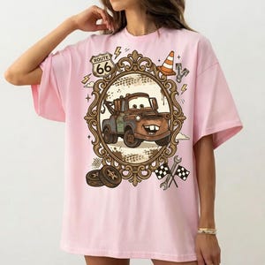 K&ouml;nnte beinhalten: Hellrosa T-Shirt mit einem Cartoon-Abschleppwagen-Design in einem verzierten Rahmen. Das Design beinhaltet ein Route 66-Schild, einen Verkehrskegel und Zielflaggen. Das T-Shirt ist hellrosa.
