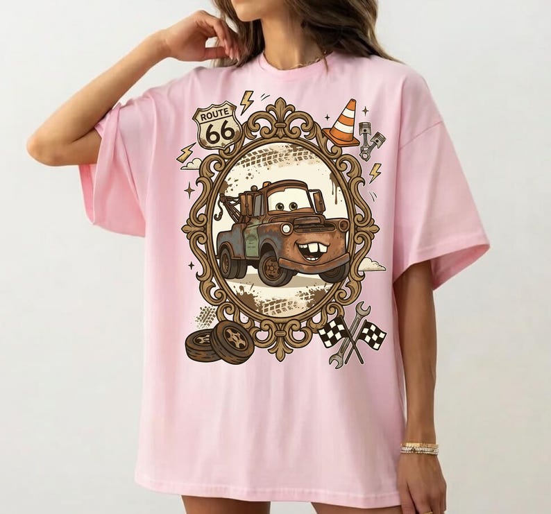 K&ouml;nnte beinhalten: Hellrosa T-Shirt mit einem Cartoon-Abschleppwagen-Design in einem verzierten Rahmen. Das Design beinhaltet ein Route 66-Schild, einen Verkehrskegel und Zielflaggen. Das T-Shirt ist hellrosa.