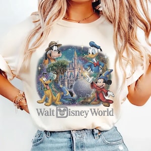 Op de afbeelding: Cr&egrave;mekleurig T-shirt met een kleurrijke afbeelding van Disney-personages, waaronder Mickey Mouse, Donald Duck en Pluto, met het kasteel van Assepoester en de tekst "Walt Disney World". Het ontwerp is geplaatst tegen een sterrenachtergrond.
