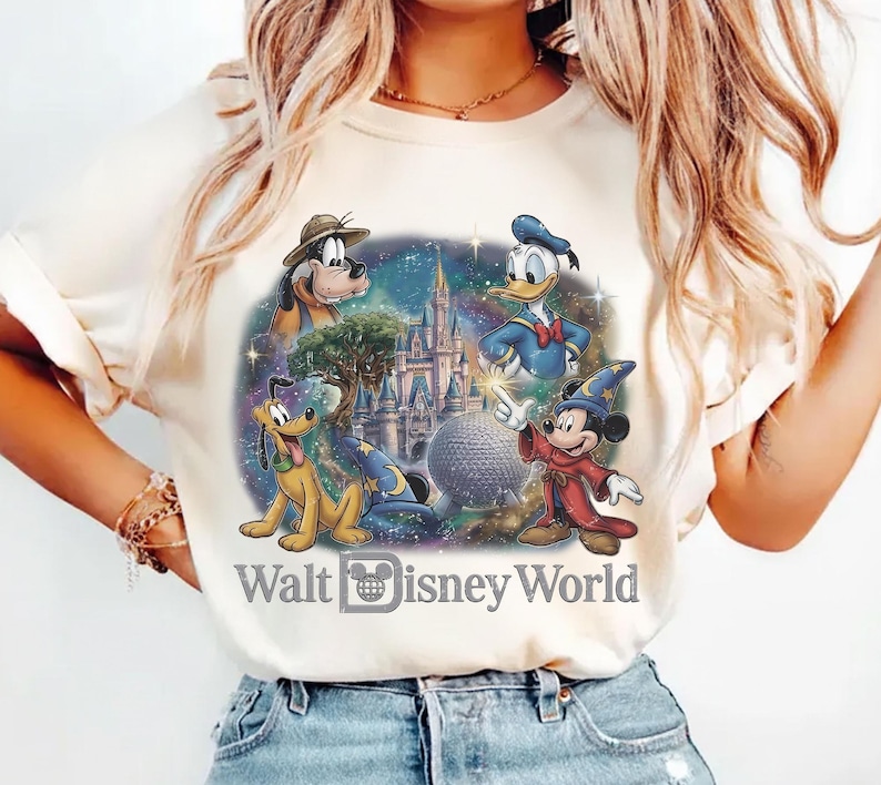 Op de afbeelding: Cr&egrave;mekleurig T-shirt met een kleurrijke afbeelding van Disney-personages, waaronder Mickey Mouse, Donald Duck en Pluto, met het kasteel van Assepoester en de tekst "Walt Disney World". Het ontwerp is geplaatst tegen een sterrenachtergrond.
