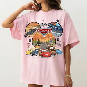 May include: Pink t-shirt featuring a graphic of the Disney Pixar Cars movie. The design includes the Cars logo, images of the characters, and racing scenes. The shirt has a crew neck and short sleeves.