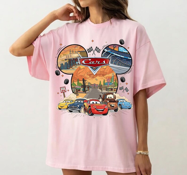 May include: Pink t-shirt featuring a graphic of the Disney Pixar Cars movie. The design includes the Cars logo, images of the characters, and racing scenes. The shirt has a crew neck and short sleeves.