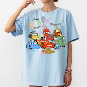May include: Light blue t-shirt featuring a colorful graphic of cartoon cars, balloons, and a birthday cake. The text "Name 7" is printed above the cars. The design includes various vehicles with party hats, creating a festive theme.