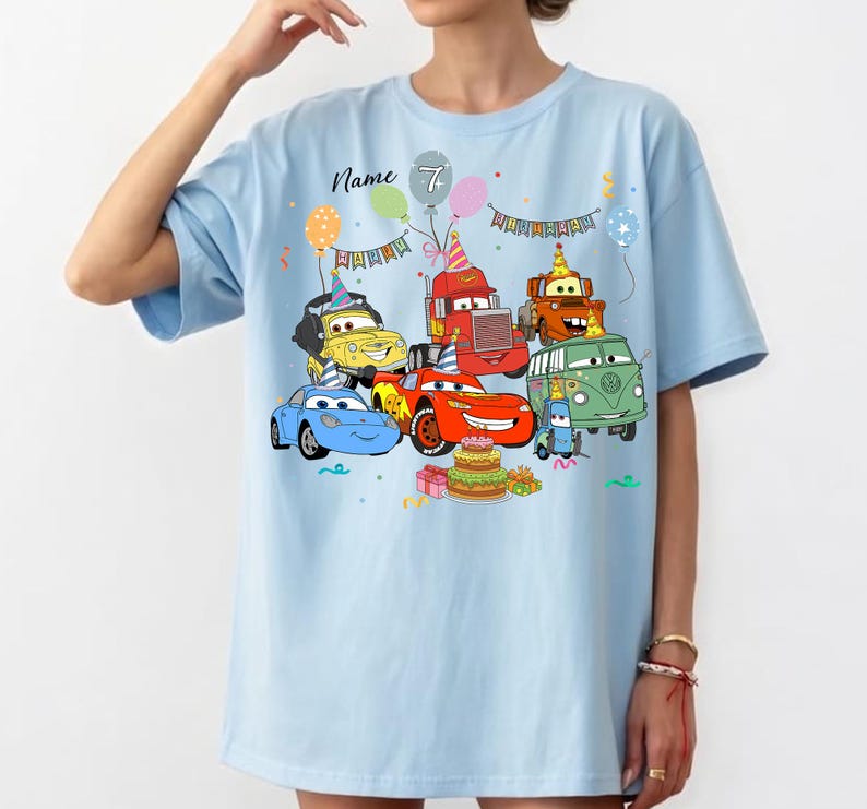 May include: Light blue t-shirt featuring a colorful graphic of cartoon cars, balloons, and a birthday cake. The text "Name 7" is printed above the cars. The design includes various vehicles with party hats, creating a festive theme.