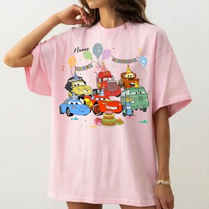 May include: Pink t-shirt featuring a colorful graphic of cartoon cars, balloons, and a birthday cake. The design includes the text "Name" and the number "7". A fun and festive design for a birthday celebration.