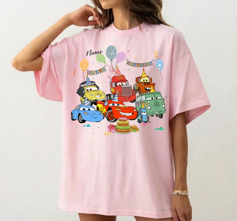 May include: Pink t-shirt featuring a colorful graphic of cartoon cars, balloons, and a birthday cake. The design includes the text "Name" and the number "7". A fun and festive design for a birthday celebration.