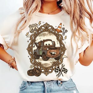 K&ouml;nnte beinhalten: Cremefarbenes T-Shirt mit einem Cartoon-Bild von Hook aus dem Film Cars, eingerahmt in einem verzierten braunen Oval. Das Design umfasst Route 66-Schilder, einen Verkehrskegel und Zielflaggen. Das Shirt wird mit einer blauen Jeans kombiniert.