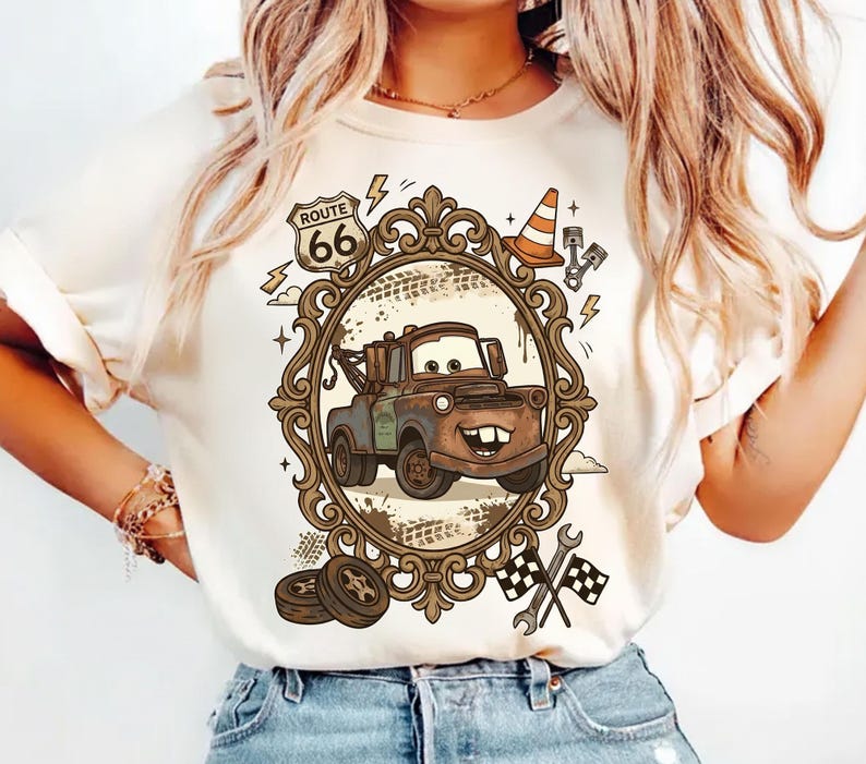 K&ouml;nnte beinhalten: Cremefarbenes T-Shirt mit einem Cartoon-Bild von Hook aus dem Film Cars, eingerahmt in einem verzierten braunen Oval. Das Design umfasst Route 66-Schilder, einen Verkehrskegel und Zielflaggen. Das Shirt wird mit einer blauen Jeans kombiniert.