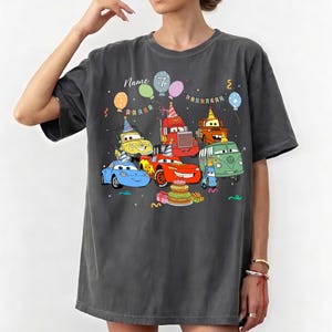 May include: Dark gray t-shirt featuring a colorful cartoon design of animated cars celebrating a birthday. The design includes a birthday cake, balloons, and the number 7. The word "Name" is also visible.