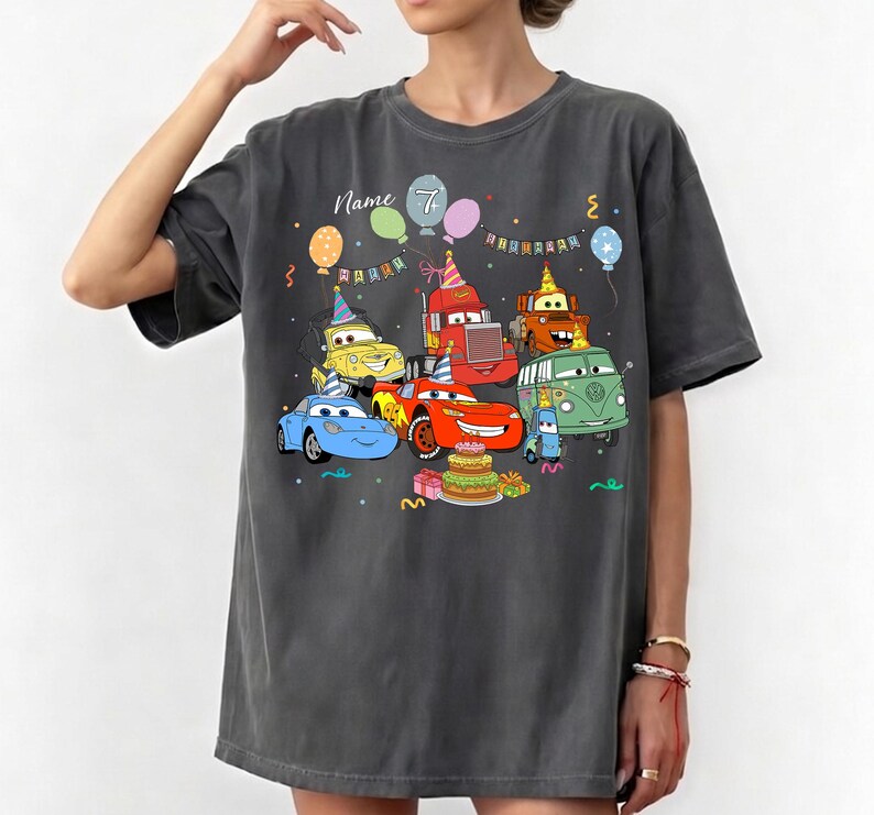 May include: Dark gray t-shirt featuring a colorful cartoon design of animated cars celebrating a birthday. The design includes a birthday cake, balloons, and the number 7. The word "Name" is also visible.
