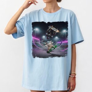 K&ouml;nnte beinhalten: Hellblaues T-Shirt mit einer Grafik von Baby Yoda, der in einem Skatepark skateboardet. Das Design zeigt einen Skateboarder, der einen Trick macht, und Baby Yoda auf einem gr&uuml;nen Skateboard. Der Hintergrund ist ein neonbeleuchteter Skatepark.