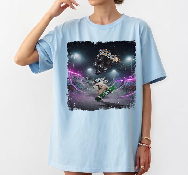 K&ouml;nnte beinhalten: Hellblaues T-Shirt mit einer Grafik von Baby Yoda, der in einem Skatepark skateboardet. Das Design zeigt einen Skateboarder, der einen Trick macht, und Baby Yoda auf einem gr&uuml;nen Skateboard. Der Hintergrund ist ein neonbeleuchteter Skatepark.