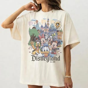 Op de afbeelding: Een off-white t-shirt met korte mouwen en een kleurrijke grafische print van Disney-figuren, waaronder Mickey Mouse, Minnie Mouse, Donald Duck, Goofy en Pluto voor een kasteel, met de tekst Disneyland Est. 1955 eronder gedrukt.