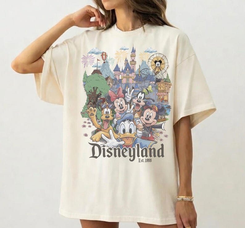 Op de afbeelding: Een off-white t-shirt met korte mouwen en een kleurrijke grafische print van Disney-figuren, waaronder Mickey Mouse, Minnie Mouse, Donald Duck, Goofy en Pluto voor een kasteel, met de tekst Disneyland Est. 1955 eronder gedrukt.
