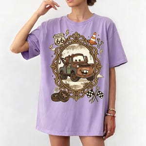 K&ouml;nnte beinhalten: Helllila T-Shirt mit einem Cartoon-Bild von Hook aus dem Film Cars, in einem verzierten Rahmen. Das Design beinhaltet Route 66-Schilder, einen Verkehrskegel und Rennflaggen. Dies ist ein Freizeitkleidungsst&uuml;ck.