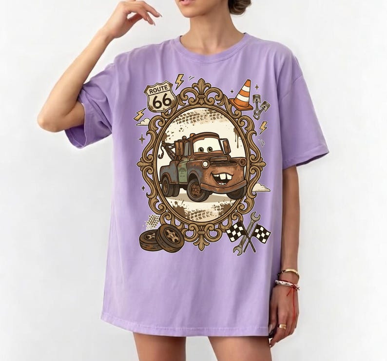K&ouml;nnte beinhalten: Helllila T-Shirt mit einem Cartoon-Bild von Hook aus dem Film Cars, in einem verzierten Rahmen. Das Design beinhaltet Route 66-Schilder, einen Verkehrskegel und Rennflaggen. Dies ist ein Freizeitkleidungsst&uuml;ck.