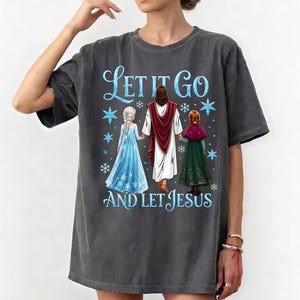 May include: Dark gray t-shirt featuring the text "Let It Go AND LET JESUS" in blue script. The graphic includes illustrations of Jesus and two characters from the movie Frozen, with snowflakes. The shirt is a casual, oversized style.
