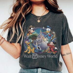 Op de afbeelding: Donkergrijs T-shirt met een kleurrijke afbeelding van Disney-personages, waaronder Mickey Mouse, Donald Duck en Pluto. Het ontwerp bevat een kasteel, een bol en de tekst "Walt Disney World".