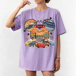 May include: A light purple t-shirt featuring a graphic of the movie Cars. The graphic includes the Cars logo, images of the characters, and scenes from the movie. The shirt has short sleeves and a crew neck.