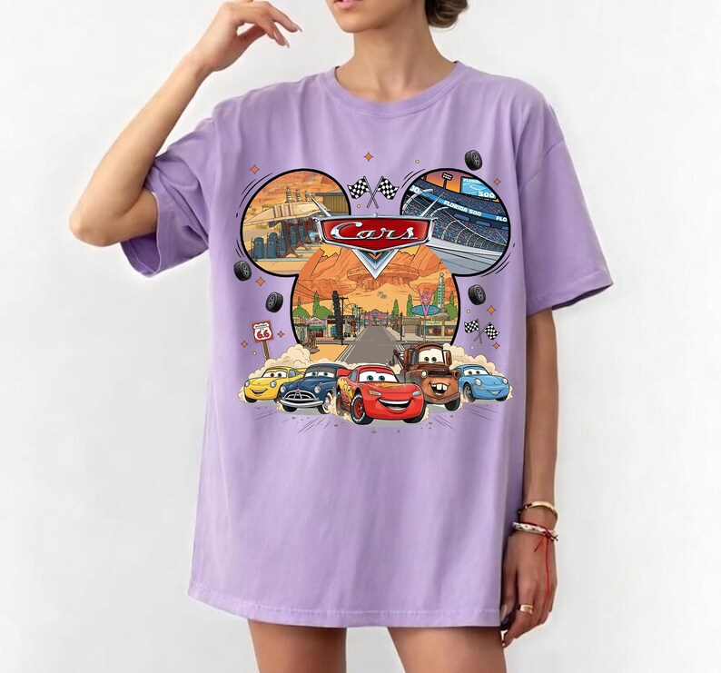 May include: A light purple t-shirt featuring a graphic of the movie Cars. The graphic includes the Cars logo, images of the characters, and scenes from the movie. The shirt has short sleeves and a crew neck.