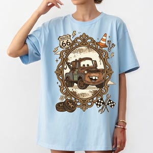 K&ouml;nnte beinhalten: Hellblaues T-Shirt mit einem Cartoon-Bild von Hook aus dem Film Cars, umrahmt von einem dekorativen Rahmen mit Route 66-Schild. Das Design umfasst Blitze, einen Verkehrskegel und Rennflaggen.