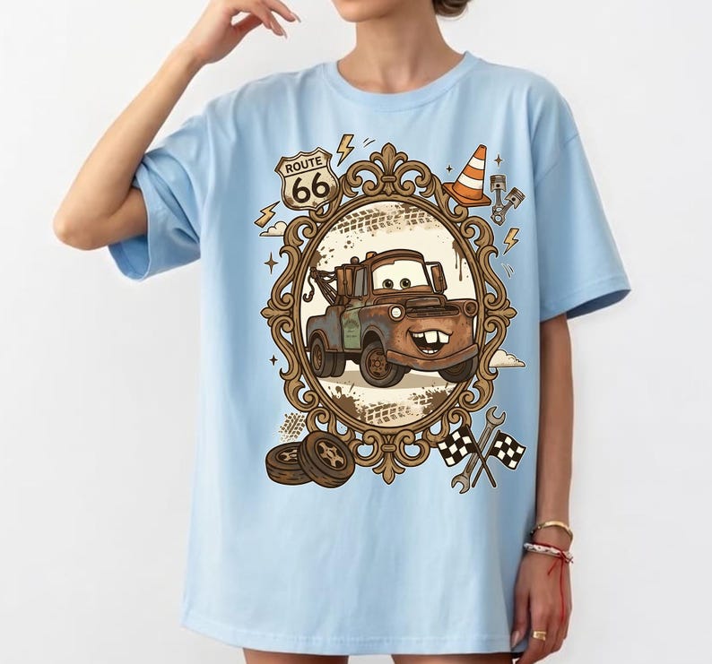 K&ouml;nnte beinhalten: Hellblaues T-Shirt mit einem Cartoon-Bild von Hook aus dem Film Cars, umrahmt von einem dekorativen Rahmen mit Route 66-Schild. Das Design umfasst Blitze, einen Verkehrskegel und Rennflaggen.