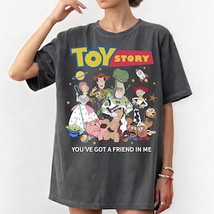 May include: Dark gray t-shirt featuring the Toy Story logo in yellow and red, with a colorful graphic of the movie's characters. The text "YOU'VE GOT A FRIEND IN ME" is printed below the graphic.