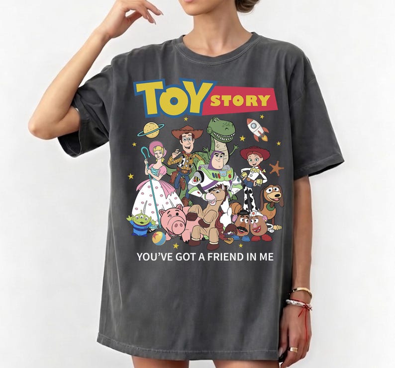 May include: Dark gray t-shirt featuring the Toy Story logo in yellow and red, with a colorful graphic of the movie's characters. The text "YOU'VE GOT A FRIEND IN ME" is printed below the graphic.