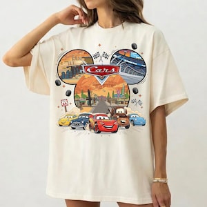 May include: Cream-colored t-shirt featuring a colorful graphic of the Disney Pixar Cars movie. The design includes the Cars logo, race cars, and Route 66 signage. The shirt has short sleeves and a relaxed fit.