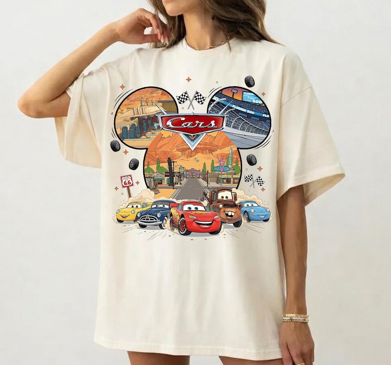 May include: Cream-colored t-shirt featuring a colorful graphic of the Disney Pixar Cars movie. The design includes the Cars logo, race cars, and Route 66 signage. The shirt has short sleeves and a relaxed fit.