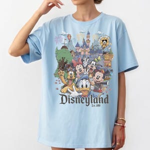 Op de afbeelding: Een lichtblauw t-shirt met een kleurrijke afbeelding van Disney-figuren waaronder Mickey Mouse, Minnie Mouse, Donald Duck, Goofy en Pluto voor een kasteel, met de tekst Disneyland Est. 1955 in donkergrijs onder de figuren gedrukt.