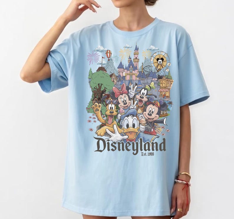 Op de afbeelding: Een lichtblauw t-shirt met een kleurrijke afbeelding van Disney-figuren waaronder Mickey Mouse, Minnie Mouse, Donald Duck, Goofy en Pluto voor een kasteel, met de tekst Disneyland Est. 1955 in donkergrijs onder de figuren gedrukt.