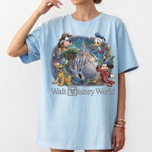 Op de afbeelding: Lichtblauw T-shirt met een kleurrijke afbeelding van Disney-personages, waaronder Mickey Mouse, Donald Duck, Pluto en Goofy. De tekst "Walt Disney World" is ook aanwezig.