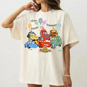 May include: Cream-colored t-shirt featuring a colorful graphic of cartoon cars, balloons, and a birthday cake. The text "Name" and the number "7" are also visible. The shirt has short sleeves and a relaxed fit.