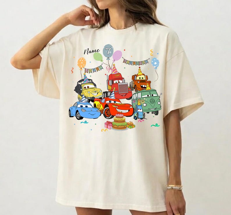 May include: Cream-colored t-shirt featuring a colorful graphic of cartoon cars, balloons, and a birthday cake. The text "Name" and the number "7" are also visible. The shirt has short sleeves and a relaxed fit.