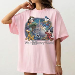 Op de afbeelding: Lichtroze T-shirt met een kleurrijke afbeelding van Disney-personages Pluto, Donald Duck, Mickey Mouse en een kasteel. De tekst "Walt Disney World" staat eronder.