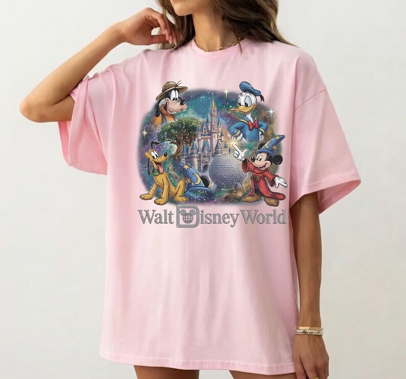 Op de afbeelding: Lichtroze T-shirt met een kleurrijke afbeelding van Disney-personages Pluto, Donald Duck, Mickey Mouse en een kasteel. De tekst "Walt Disney World" staat eronder.