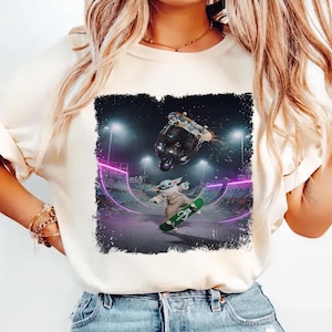 K&ouml;nnte beinhalten: Cremefarbenes T-Shirt mit einer Grafik von Baby Yoda beim Skateboarden in einem Skatepark. Das Design umfasst einen dunklen Hintergrund mit lila Neonlichtern und einen Skateboarder in der Luft.