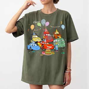 May include: Olive green t-shirt featuring a colorful cartoon car design with balloons, a birthday cake, and the number 7. The text "Name" is above the car graphic. The shirt is a casual, oversized fit.