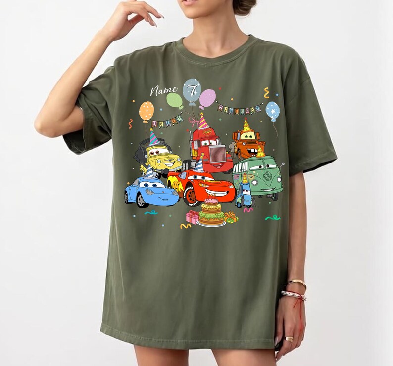 May include: Olive green t-shirt featuring a colorful cartoon car design with balloons, a birthday cake, and the number 7. The text "Name" is above the car graphic. The shirt is a casual, oversized fit.