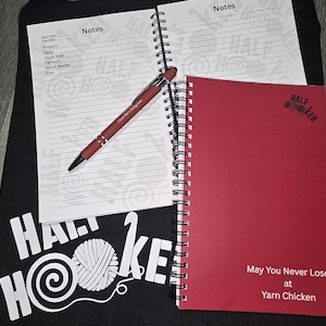 May include: A red spiral-bound notebook and a lined notepad with a pen, and a black t-shirt with the words "Half Hooked" and a yarn ball graphic. The notebook has the text "May You Never Lose at Yarn Chicken".