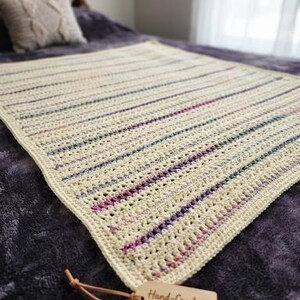 May include: A rectangular crochet blanket featuring a cream-colored base with horizontal stripes in shades of purple, pink, and blue. A light brown wooden tag is attached with a leather cord, displaying the text Hand-Crocheted with Love.