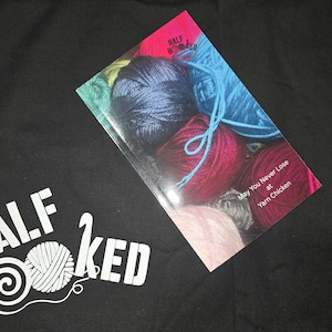 May include: Black fabric with the white text "HALF HOOKED" and a yarn ball graphic. A colorful card with yarn balls and the text "May You Never Lose at Yarn Chicken" is on top of the fabric.