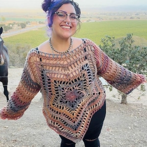 May include: A long-sleeved crochet sweater featuring a large, multi-pointed star pattern in variegated earth tones including brown, rust, gray, and muted purple. The garment has an open-knit construction, a wide neckline, and a pointed hemline.
