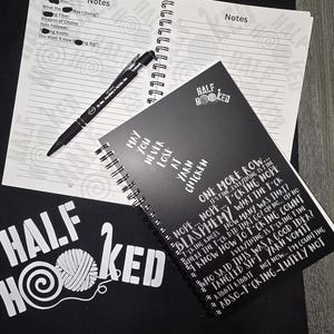 May include: A black tote bag with the words "HALF HOOKED" in white, along with a yarn ball and crochet hook design. A spiral-bound notebook with handwritten notes and a black pen are on top. Another notebook with humorous text is also visible.